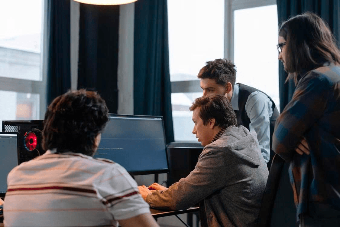Four men gather around a desktop monitor in an office, one typing while the others look on.