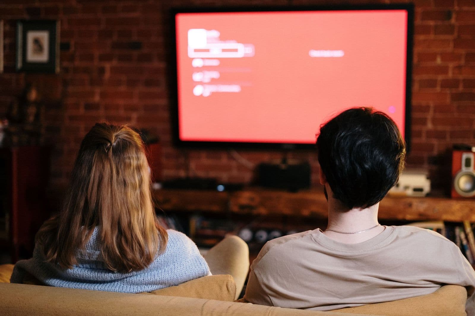 Two people seen from behind on a sofa, facing a wall-mounted television showing a red home screen in a brick-walled living room.