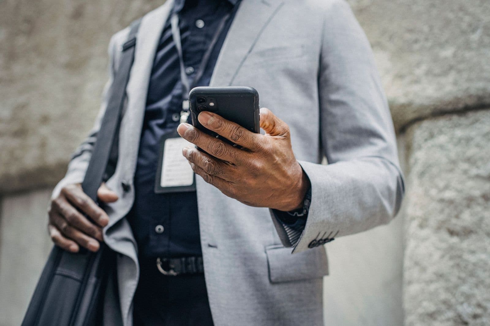 Close view of a man in a grey blazer and dark shirt with a lanyard holding a smartphone, a bag strap over his shoulder