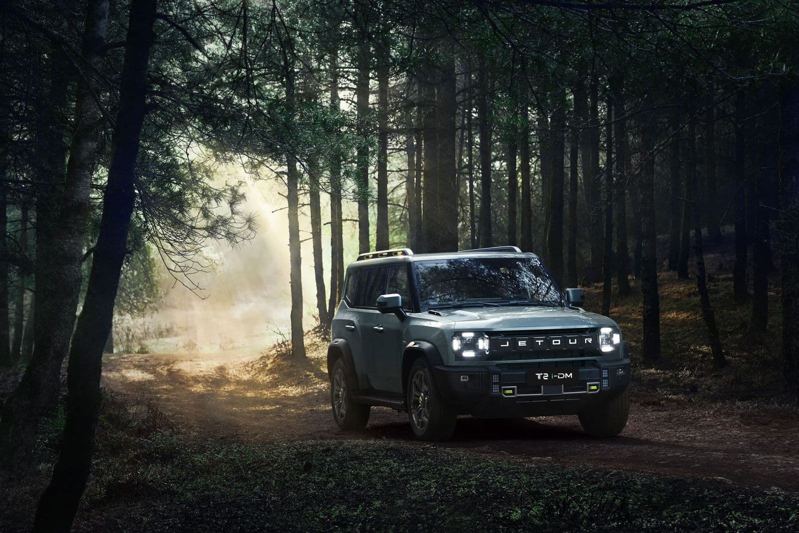 A grey Jetour T2 i-DM SUV with its headlights on drives along a dirt trail through a misty pine forest