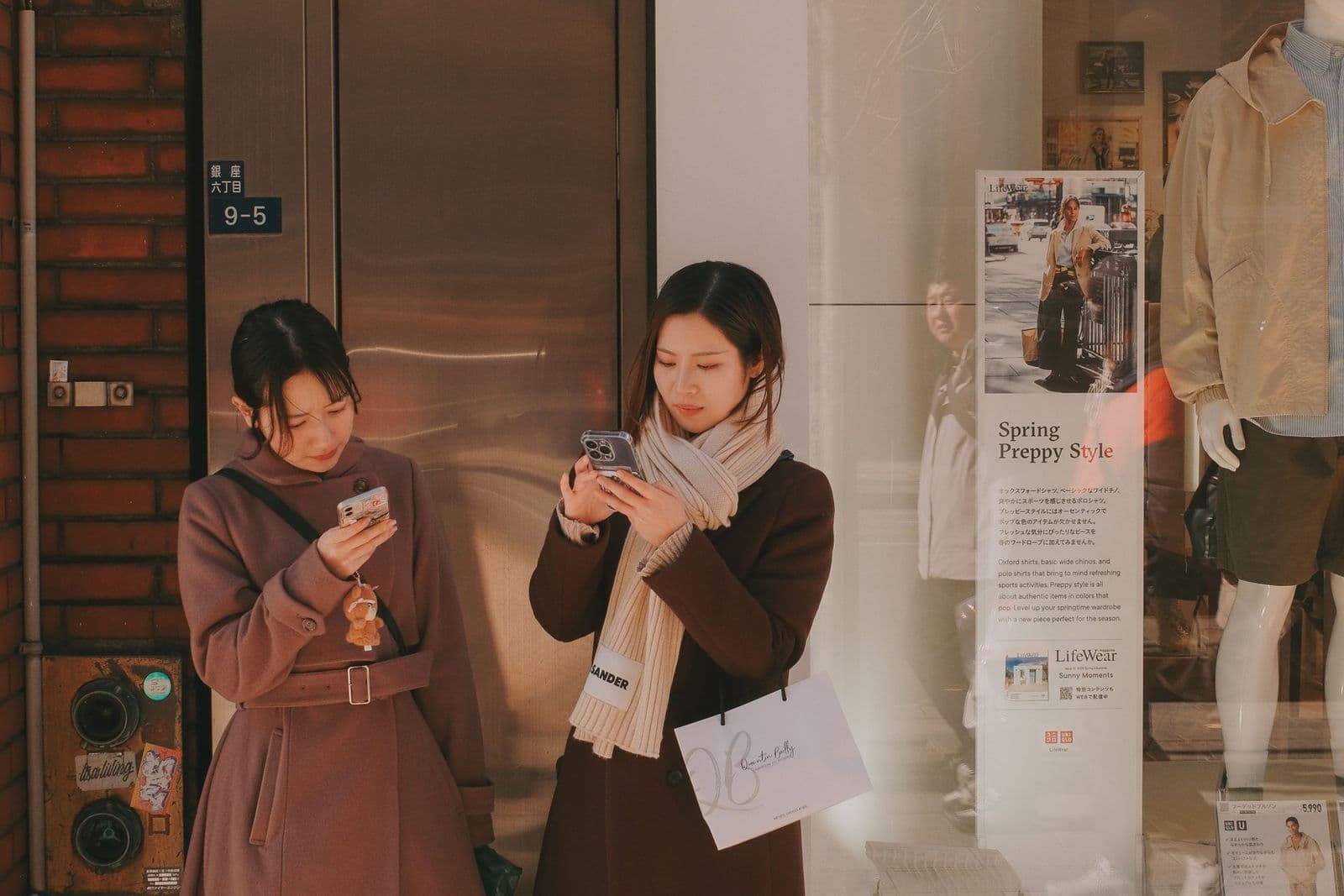 Two young women in coats stand on a city pavement looking at their smartphones outside a shop window